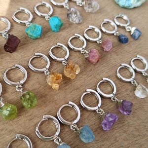 Puede incluir: Pendientes de aro plateados, cada uno adornado con una piedra preciosa colorida única. Las piedras preciosas varían en color, incluyendo turquesa, púrpura, amarillo, verde y transparente. Los pendientes se exhiben sobre una superficie de madera.
