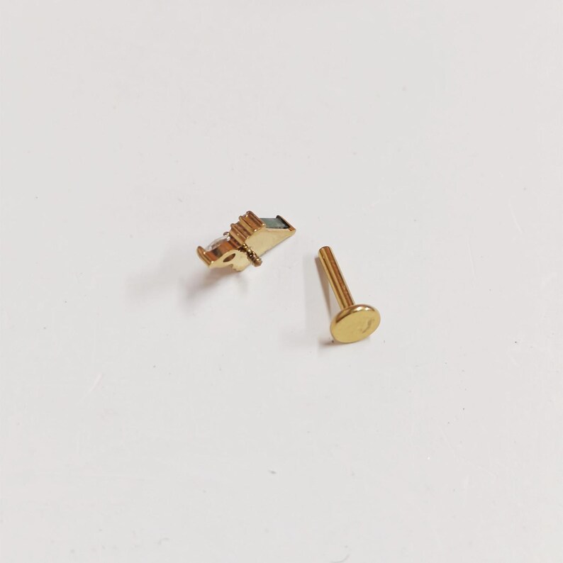 May include: Two gold-colored body jewelry pieces. One piece is a flat-backed stud with a round, flat head. The other piece is a small, rectangular shape with a small, rectangular, dark-colored stone.