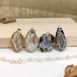 May include: Four agate pendant charms with gold-plated edges. Each pendant features a unique natural agate pattern with a hollow centre. The pendants are displayed on a wooden surface with a pearl necklace.