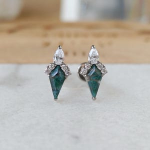 May include: A pair of stud earrings featuring a geometric design. Each earring has a teardrop-shaped, clear gemstone at the top, a cluster of small, clear stones, and a green and blue, diamond-shaped stone. The earrings are set on a silver post.