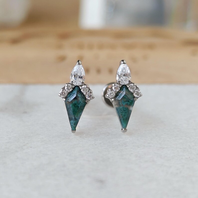 May include: A pair of stud earrings featuring a geometric design. Each earring has a teardrop-shaped, clear gemstone at the top, a cluster of small, clear stones, and a green and blue, diamond-shaped stone. The earrings are set on a silver post.
