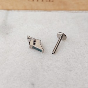 May include: A silver-colored lip piercing stud with a small, angular design and a teal-colored accent. The stud is next to a separate, straight post with a flat, round top, designed for insertion into the piercing.