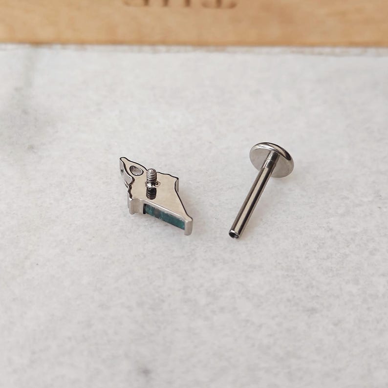 May include: A silver-colored lip piercing stud with a small, angular design and a teal-colored accent. The stud is next to a separate, straight post with a flat, round top, designed for insertion into the piercing.