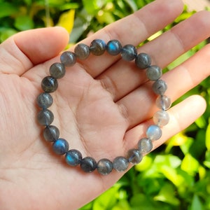 May include: A dark blue labradorite gemstone bracelet with iridescent blue and green flashes. The bracelet is made of round beads and is shown on a hand.
