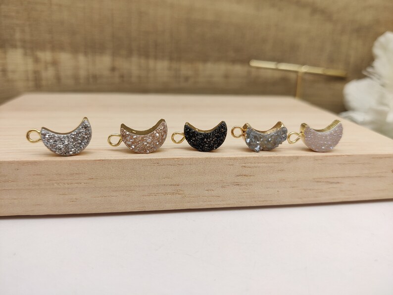 May include: Five gold-toned crescent moon charms with different colored glitter finishes. The charms are arranged in a row on a wooden surface. The colors are silver, gold, black, blue, and white.
