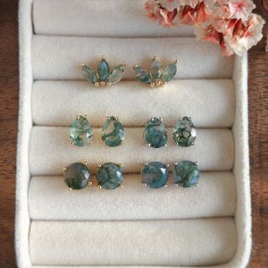 May include: Three pairs of stud earrings featuring moss agate stones in gold and silver settings. The top pair has a three-stone leaf shape, the middle pair is pear-shaped, and the bottom pair is round. Each stone displays unique green inclusions.
