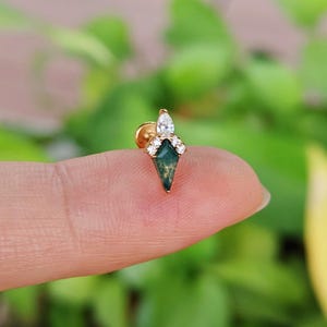 May include: A gold-colored stud earring with a green, diamond-shaped stone. The earring is adorned with small, clear crystals and a larger, teardrop-shaped crystal at the top. The earring is held on a finger against a blurred green background.