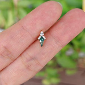May include: A small, gold-toned earring with a geometric, emerald-green gemstone at its center. The earring is adorned with a cluster of small, clear gemstones and a larger, pear-shaped stone at the top. The design is delicate and elegant.