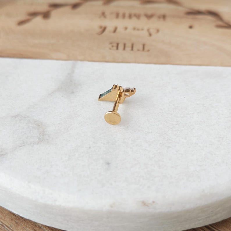 May include: A gold-colored stud earring with a small, geometric design featuring a green and clear gemstone. The earring has a flat, round backing and a decorative top.