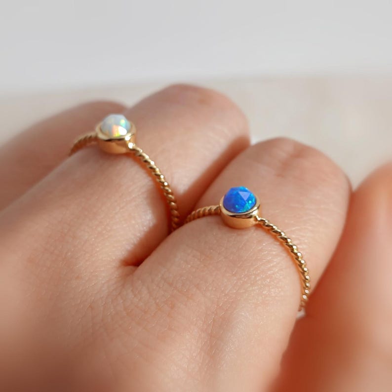 Puede incluir: Dos anillos de oro con bandas trenzadas, cada uno con una peque&ntilde;a piedra preciosa redonda facetada. Un anillo tiene una piedra blanca iridiscente, mientras que el otro tiene una piedra azul vibrante, ambos en engastes de bisel de oro simples.