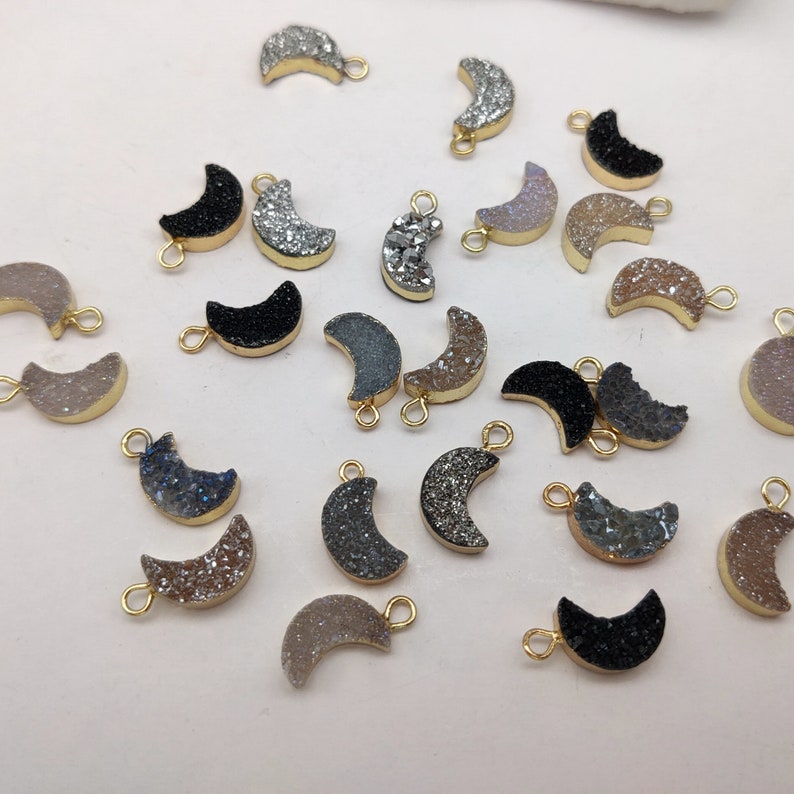 May include: A collection of 20 gold-plated crescent moon charms with various gemstone finishes, including black, white, grey, and brown. The charms have a hole at the top for attaching to jewellery.