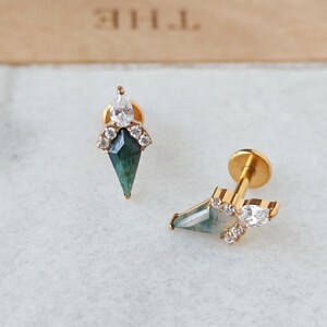 May include: Two gold-colored stud earrings. Each earring features a teardrop-shaped white gem at the top, a cluster of small clear gems, and a green, diamond-shaped gem. The earrings are on a white surface.