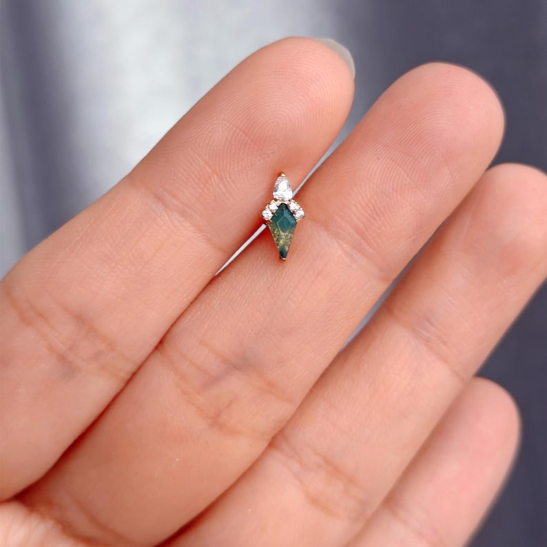 May include: A small, gold-toned earring with a diamond-shaped, green gemstone at its center. The earring is adorned with small, clear gemstones at the top. The design is delicate and elegant, suitable for ear piercings.