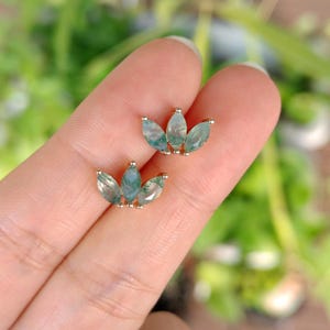 May include: A pair of gold-toned stud earrings, each featuring a cluster of three marquise-cut gemstones. The gemstones have a translucent, pale green hue with darker green inclusions, resembling a floral design.