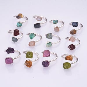 Pode incluir: Uma coleção de doze anéis abertos de metal prateado, cada um com duas pedras preciosas brutas e não lapidadas em várias cores, incluindo roxo, verde, azul, rosa, castanho e transparente, dispostos numa grelha sobre um fundo branco.