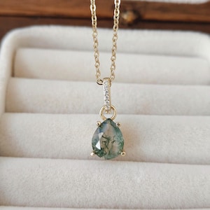 May include: A gold-colored necklace with a teardrop-shaped pendant. The pendant is a light green color with darker green inclusions. The necklace chain is delicate, and the pendant is set with small, clear stones.