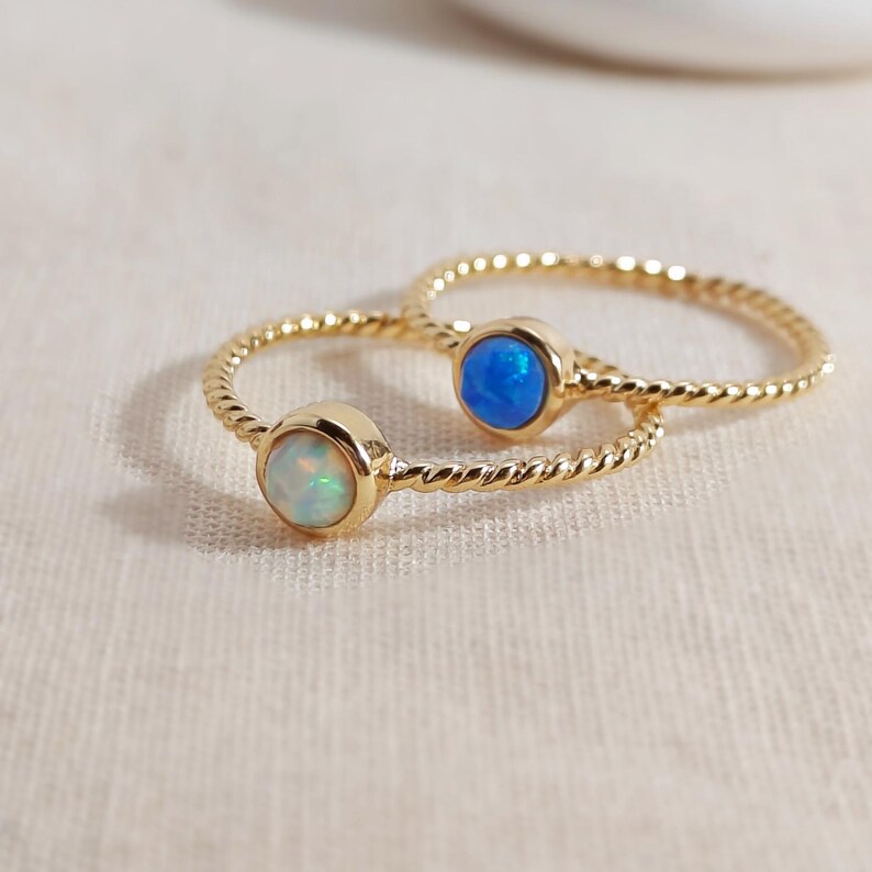 Puede incluir: Dos anillos de tono dorado con dise&ntilde;o de banda trenzada. Un anillo tiene una piedra redonda blanca iridiscente similar al &oacute;palo, mientras que el otro tiene una piedra redonda de color azul vibrante. Ambos anillos tienen un acabado met&aacute;lico pulido.