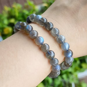May include: Two beaded bracelets made of iridescent blue-grey labradorite stones. The bracelets are stacked on a person's wrist.