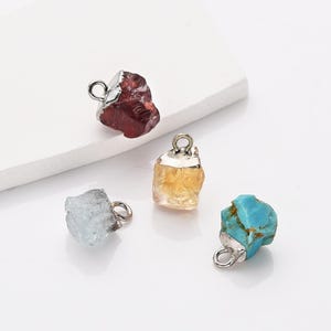 May include: Four raw gemstone pendants with silver jump rings. The pendants are red, yellow, blue, and light blue.