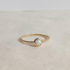 Puede incluir: Un anillo de oro con una banda de estilo cuerda trenzada y una piedra preciosa de &oacute;palo redonda engastada en bisel. El &oacute;palo muestra destellos iridiscentes de color, y la banda de metal dorado pulido crea una forma circular continua alrededor.