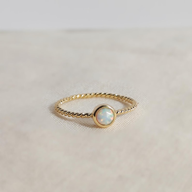 Puede incluir: Un anillo de oro con una banda de estilo cuerda trenzada y una piedra preciosa de &oacute;palo redonda engastada en bisel. El &oacute;palo muestra destellos iridiscentes de color, y la banda de metal dorado pulido crea una forma circular continua alrededor.