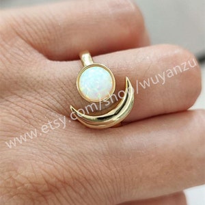 Peut inclure: Bague en or avec une pierre d'opale blanche sertie dans un motif de croissant de lune. La bague est portée sur un doigt.