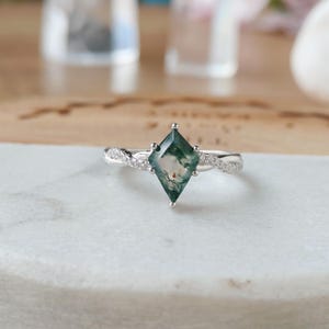 Puede incluir: Un anillo de plata con una gema de ágata musgosa verde en forma de diamante. La banda tiene un diseño retorcido con pequeñas piedras de acento claras. El anillo está sobre una superficie de mármol blanco.
