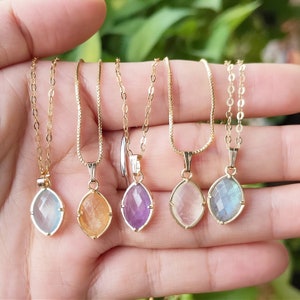 May include: Five gold necklaces with different colored gemstone pendants. The pendants are faceted and have a teardrop shape. The necklaces are on a hand, and the gemstones are amethyst, labradorite, clear, orange, and blue.