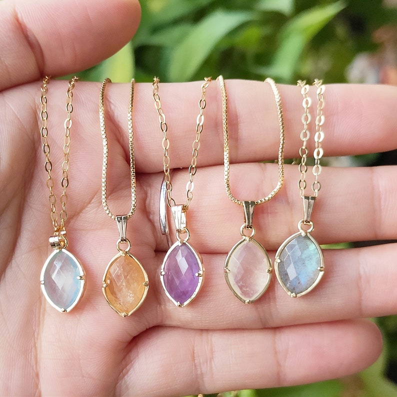 May include: Five gold necklaces with different colored gemstone pendants. The pendants are faceted and have a teardrop shape. The necklaces are on a hand, and the gemstones are amethyst, labradorite, clear, orange, and blue.