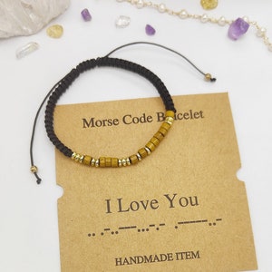 Puede incluir: Un brazalete de cuerda negro con cuentas doradas que deletrean "Te amo" en código Morse en una tarjeta marrón con el texto "Brazalete de código Morse" y "Hecho a mano".