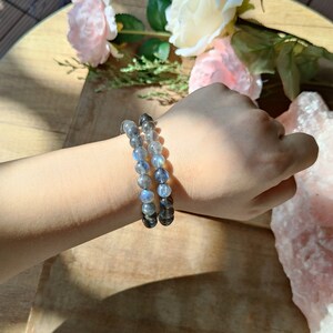 May include: Two beaded bracelets made of labradorite stones. The bracelets are stacked on a person's wrist. The stones are a mix of dark gray and iridescent blue.
