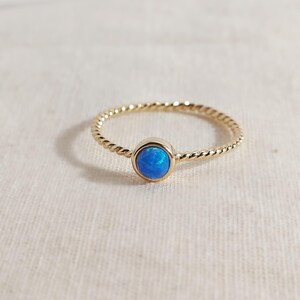 Puede incluir: Un anillo de oro con una banda fina y trenzada y una piedra de &oacute;palo azul redonda y facetada engastada en un bisel de oro liso. La vibrante piedra preciosa azul muestra sutiles destellos iridiscentes, contrastando con la banda de oro amarillo pulido.