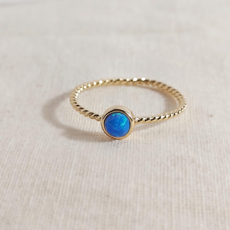Puede incluir: Un anillo de oro con una banda fina y trenzada y una piedra de &oacute;palo azul redonda y facetada engastada en un bisel de oro liso. La vibrante piedra preciosa azul muestra sutiles destellos iridiscentes, contrastando con la banda de oro amarillo pulido.