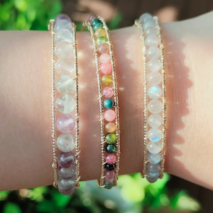 May include: Three gold wire wrapped bracelets with different colored gemstone beads. The bracelets feature pink, green, and white beads.