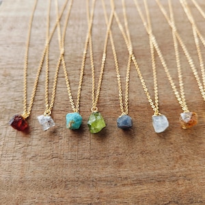 Op de afbeelding: Een verzameling goudkleurige kettingkettingen, elk met een andere ruwe edelsteen hanger. De edelstenen variëren in kleur, waaronder rood, helder, turquoise, groen, blauw en oranje. De kettingen worden gepresenteerd op een houten oppervlak.