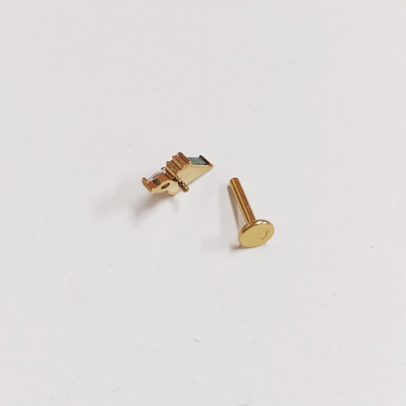 May include: Two gold-colored body jewelry pieces. One piece features a small, rectangular design with a dark accent, and the other is a straight post with a flat, round end. The jewelry is set against a white background.