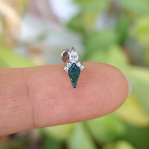 May include: A silver-toned earring with a dark teal, kite-shaped center stone. The earring is adorned with small, clear gemstones and a pear-shaped stone at the top. The earring is held on a finger against a blurred green background.