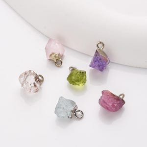 May include: Six different colored gemstone charms with silver jump rings. The charms are shaped like rough, uncut crystals. The colors are pink, clear, green, purple, and red.