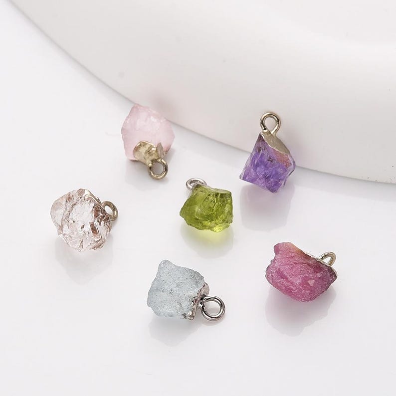 May include: Six different colored gemstone charms with silver jump rings. The charms are shaped like rough, uncut crystals. The colors are pink, clear, green, purple, and red.