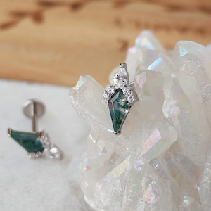 May include: A pair of silver earrings with a geometric design. Each earring features a green and white stone in a kite shape, surrounded by small, clear crystals. The earrings are displayed on a white, crystalline surface.