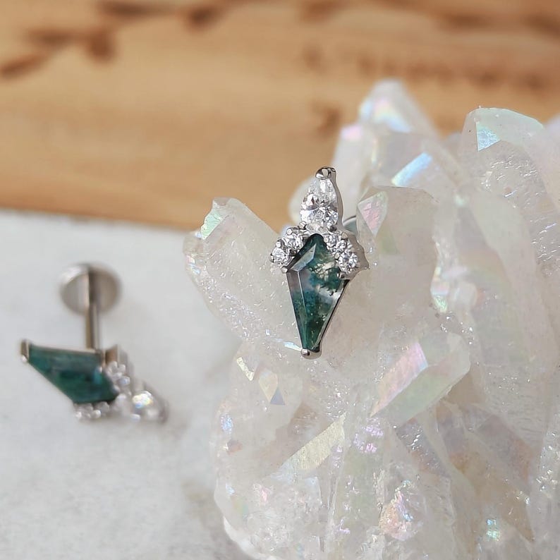 May include: A pair of silver earrings with a geometric design. Each earring features a green and white stone in a kite shape, surrounded by small, clear crystals. The earrings are displayed on a white, crystalline surface.