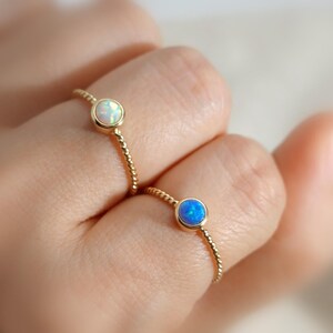 Puede incluir: Dos anillos de oro con bandas trenzadas, cada uno con una piedra preciosa redonda engastada en bisel. Un anillo tiene un &oacute;palo blanco iridiscente, mientras que el otro tiene un &oacute;palo azul vibrante. Los anillos se llevan en dos dedos separados.