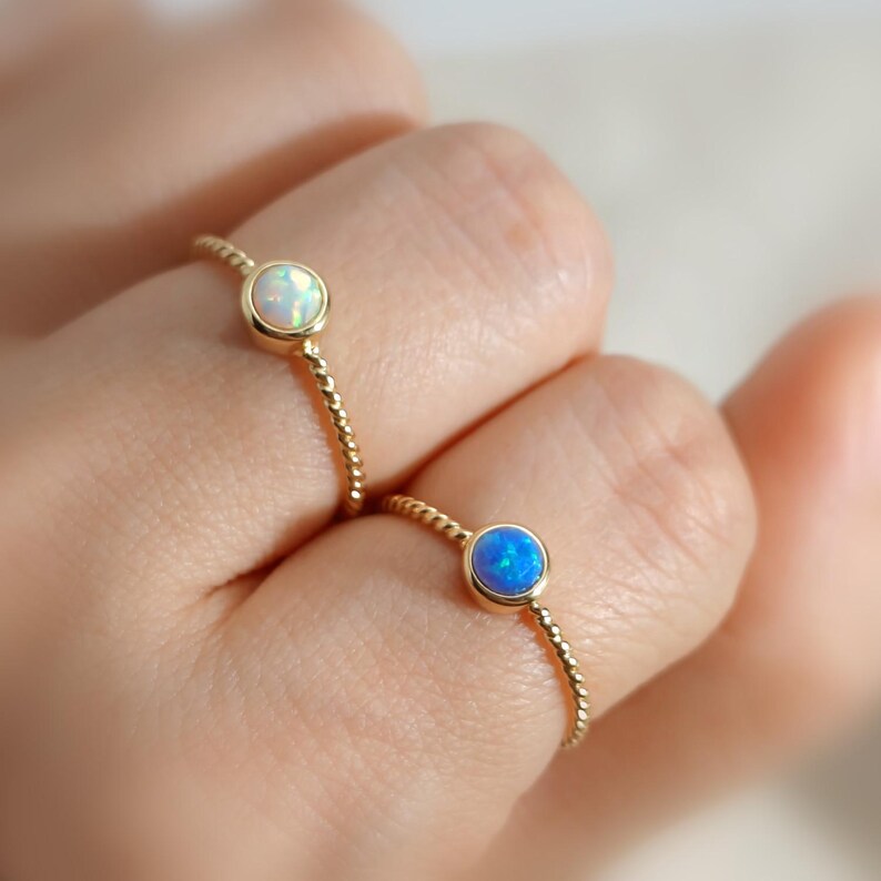 Puede incluir: Dos anillos de oro con bandas trenzadas, cada uno con una piedra preciosa redonda engastada en bisel. Un anillo tiene un &oacute;palo blanco iridiscente, mientras que el otro tiene un &oacute;palo azul vibrante. Los anillos se llevan en dos dedos separados.