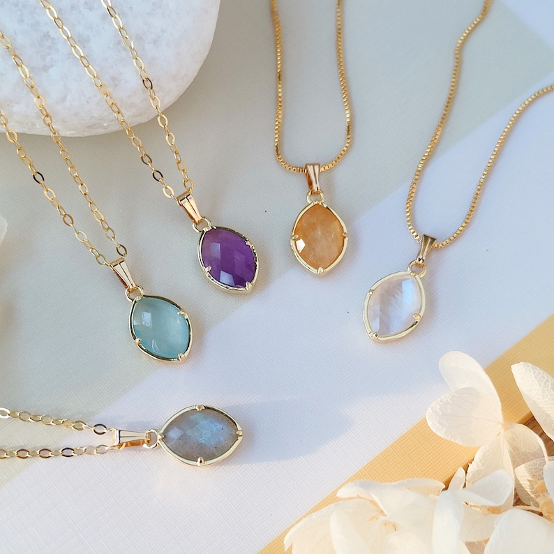 May include: Five gold necklaces with different colored gemstone pendants. The pendants are all oval shaped and have a gold setting. The gemstones are amethyst, aquamarine, moonstone, labradorite, and citrine.