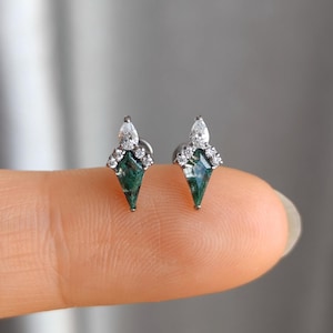 May include: A pair of stud earrings featuring a green gemstone in a kite shape, topped with a pear-shaped clear stone and surrounded by small, clear accent stones. The earrings are set in a silver metal.