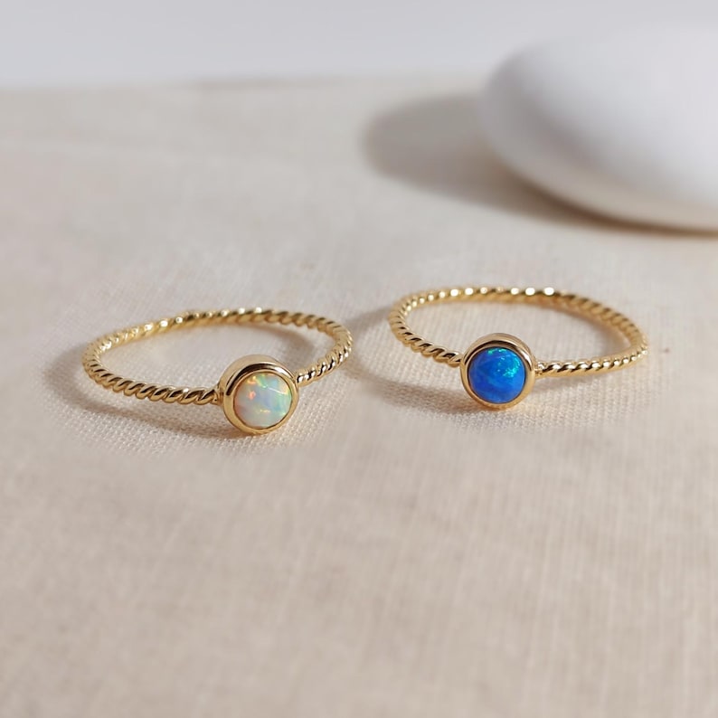 Puede incluir: Dos anillos de oro con dise&ntilde;o de banda trenzada. Un anillo presenta una piedra de &oacute;palo blanca iridiscente redonda, mientras que el otro anillo presenta una piedra de &oacute;palo azul vibrante redonda. Ambas piedras est&aacute;n en un engaste de oro liso.
