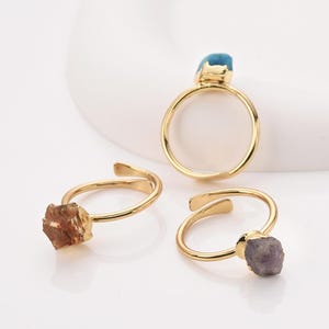 May include: Three gold-colored rings with raw gemstone settings. One ring features a light blue stone, another a brown stone, and the third a purple stone. The rings are displayed on a white surface.