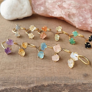 May include: A collection of gold-toned rings, each featuring a unique raw gemstone. The gemstones vary in color, including purple, yellow, orange, blue, green, and pink. The rings are displayed on a wooden surface.