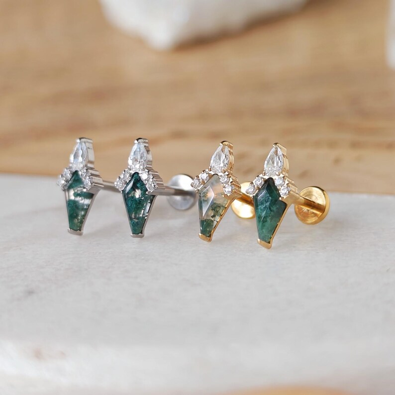 May include: Close-up of three stud earrings. Each earring features a green gemstone in a geometric shape, accented with clear crystals and a pear-shaped stone at the top. The earrings are set in silver and gold tones.