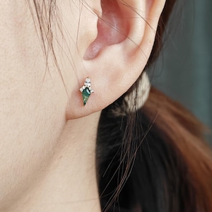 May include: A gold-colored stud earring with a green, diamond-shaped stone and small white accent stones. The earring is worn on an ear, showcasing its design and size. The earring's design is geometric and elegant.